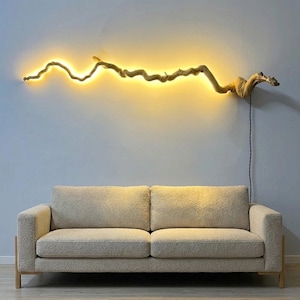 Puede incluir: Una lámpara de pared hecha con un largo trozo de madera flotante sinuosa. La madera está iluminada desde atrás por una tira de LED de color amarillo cálido, proyectando un brillo suave en la pared. Un cable negro cuelga hacia abajo.