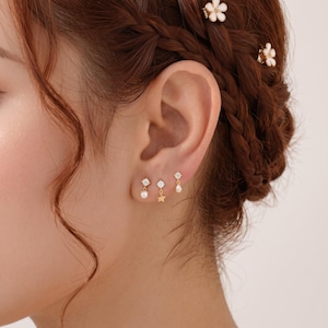 May include: Three gold-tone stud earrings with dangling charms worn in an earlobe. The first earring features a small white pearl, the second a gold star, and the third a white teardrop-shaped charm. Each stud is topped with a clear, round crystal.