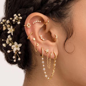 Peut inclure: Une collection de boucles d'oreilles dorées comprenant plusieurs anneaux, clous et chaînes pendantes ornées de pierres claires. Les bijoux présentent des motifs d'étoiles, de lunes et de cercles disposés sur le lobe et le cartilage, avec des fleurs blanches.