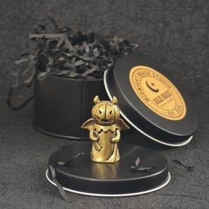 May include: A small brass figurine shaped like a pumpkin-headed creature with wings, standing in front of a black circular metal tin. The tin lid features a gold circular label with the text Midnight Brass Studio, Solid Brass, Est. 2024.