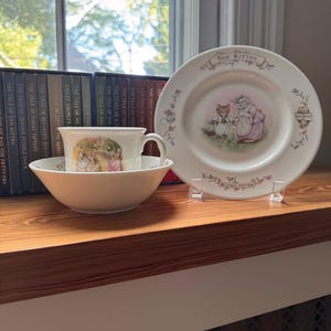 May include: A white ceramic cup, bowl, and plate set featuring Beatrix Potter illustrations. The plate displays Tom Kitten with floral accents and text. The cup and bowl show a scene with a kitten and a duck. The items are arranged on a wooden surface.