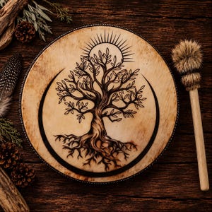 May include: A round drum with a light tan surface featuring a black pyrography design of a tree with exposed roots, a sunburst above, and crescent moon shapes on the sides. The drum is accompanied by a wooden mallet with a fluffy, light brown fur head.