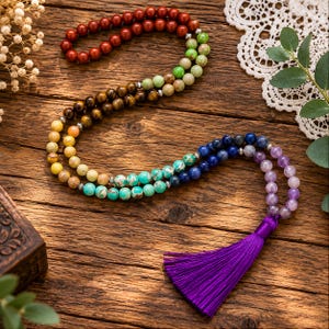 May include: A long beaded necklace featuring a gradient of round gemstone beads in shades of red, green, brown, yellow, turquoise, blue, and purple, accented with small silver-toned spacers and finished with a vibrant purple fabric tassel.