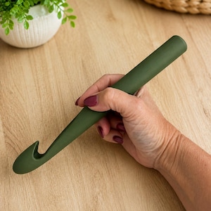 Può includere: Un uncinetto spesso di colore verde oliva opaco, con un manico cilindrico liscio e una punta curva progettata per lavorare il filato. L'attrezzo presenta un colore solido e uniforme su tutta la lunghezza, offrendo una presa salda.