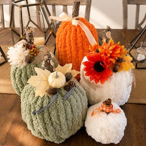 May include: A collection of five decorative pumpkins made from textured yarn in orange, sage green, and white. The pumpkins are adorned with natural elements including wooden stems, faux flowers, maple leaves, acorns, and small ribbons.