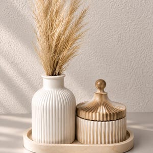 May include: A white ribbed ceramic vase containing dried beige pampas grass, a matching white ribbed lidded jar with a gold-toned finish on the lid, and a smooth oval tray. The set features a consistent vertical fluted texture throughout.