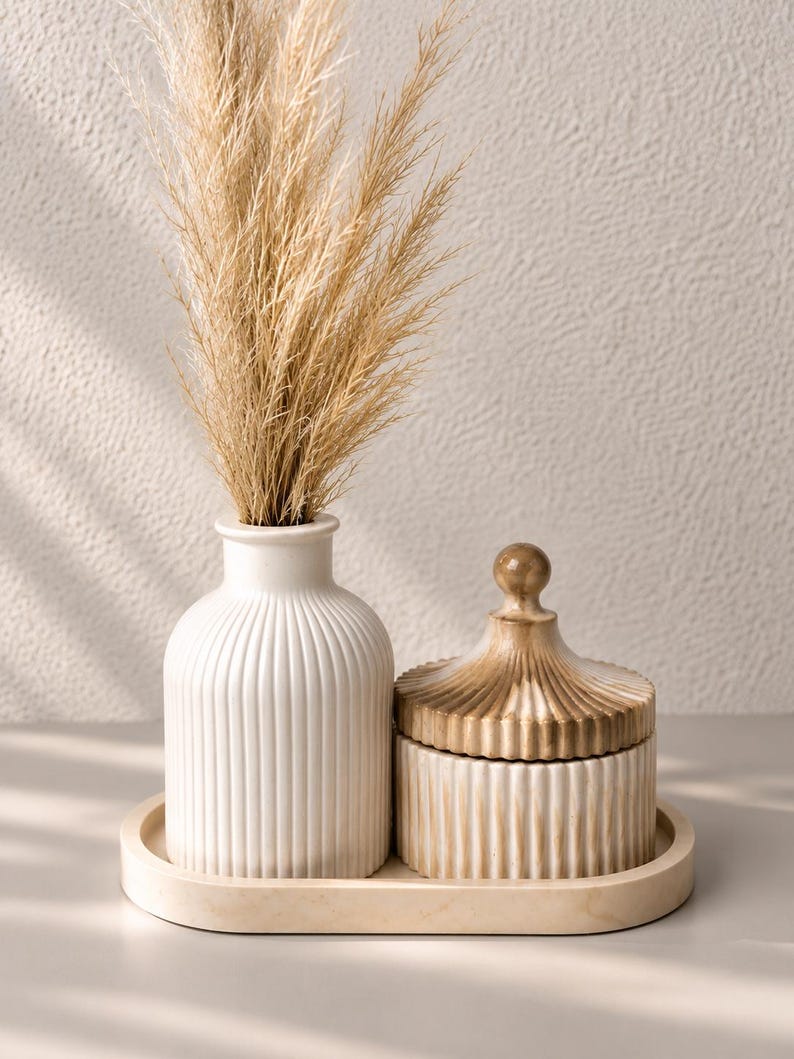 May include: A white ribbed ceramic vase containing dried beige pampas grass, a matching white ribbed lidded jar with a gold-toned finish on the lid, and a smooth oval tray. The set features a consistent vertical fluted texture throughout.