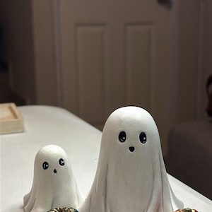 May include: Two white ghost figurines with black painted eyes and mouths, one larger than the other. A small teal pumpkin with gold accents sits to the left, and a small orange pumpkin with gold accents sits to the right of the ghosts.