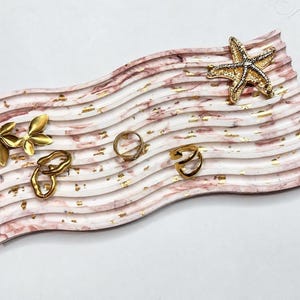 May include: A rectangular, wavy-edged tray featuring a pink and white marbled pattern with gold leaf accents. Resting on the tray are a gold starfish-shaped object, a gold leaf-shaped pin, and two gold-toned metal rings.