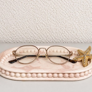 May include: An oval-shaped, light pink marble-patterned tray with a beaded edge, holding a pair of round-framed glasses with thin gold metal temples and a gold-toned, textured starfish ornament resting on the right side of the tray surface.
