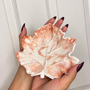 May include: A leaf-shaped trinket dish featuring a marbled pattern of white and burnt orange colors. The dish has a textured surface with raised veins that mimic the natural appearance of a leaf, with a glossy finish throughout.