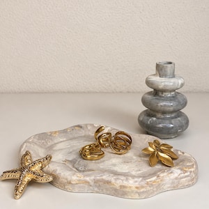 May include: A marbled beige and white oval tray holding several gold-toned rings, a gold-toned starfish ornament, and a gold-toned leaf-shaped brooch, accompanied by a small, stacked grey and white marble-patterned vase.