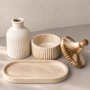 May include: A set of cream-colored home decor items including a ribbed vase, a round lidded container with a marbled finish, and an oval tray. The lid of the container is detached, showing a brown and cream marbled pattern with a small knob handle.
