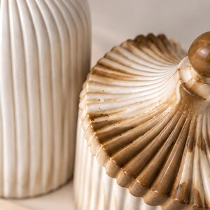 May include: A ceramic jar with a lid and a matching tall cylindrical container, both featuring a vertical ribbed texture. The pieces are finished in a cream-colored glaze with brown shading concentrated along the edges and the lid's finial.