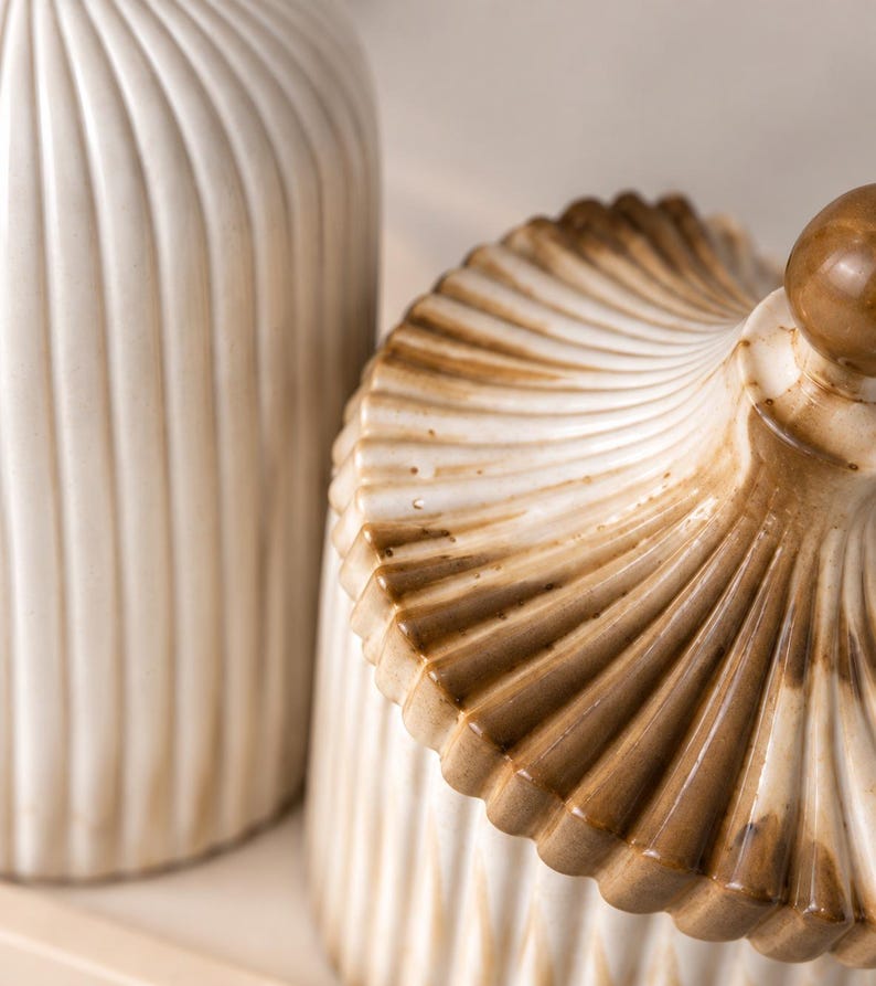 May include: A ceramic jar with a lid and a matching tall cylindrical container, both featuring a vertical ribbed texture. The pieces are finished in a cream-colored glaze with brown shading concentrated along the edges and the lid's finial.