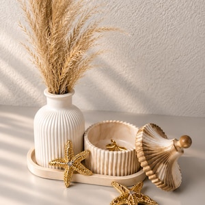 May include: A white ribbed vase containing dried beige grass, a matching lidded jar with a marble-like finish, and two gold-toned starfish ornaments arranged on an oval tray. The jar lid is placed beside the container, revealing a small gold object.