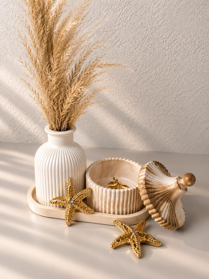 May include: A white ribbed vase containing dried beige grass, a matching lidded jar with a marble-like finish, and two gold-toned starfish ornaments arranged on an oval tray. The jar lid is placed beside the container, revealing a small gold object.