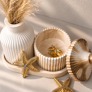 May include: A white ribbed vase, a matching lidded jar with a marbled finish, and two gold-toned starfish ornaments arranged on an oval tray. The jar contains a small gold-toned floral accent, and the vase holds a bundle of dried beige grass.