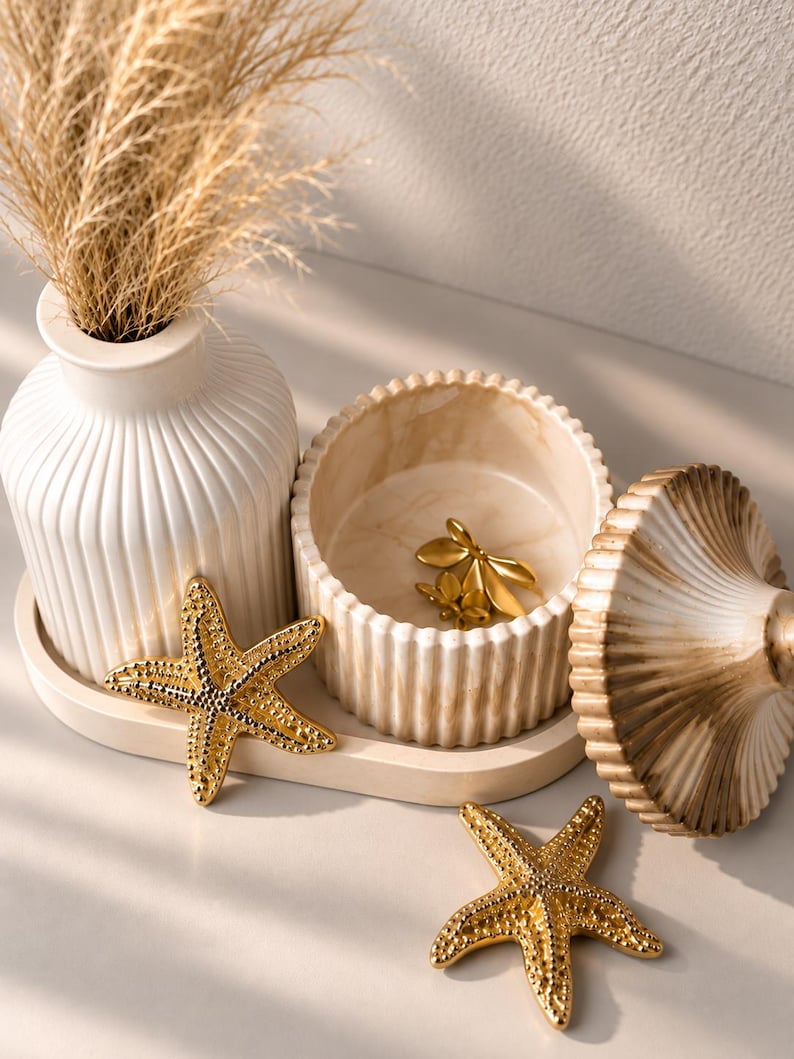 May include: A white ribbed vase, a matching lidded jar with a marbled finish, and two gold-toned starfish ornaments arranged on an oval tray. The jar contains a small gold-toned floral accent, and the vase holds a bundle of dried beige grass.