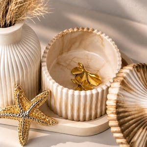 May include: A round, ribbed, cream-colored container with a marble-like finish, containing two gold-toned leaf-shaped ornaments. Beside it sits a gold-toned starfish, a matching ribbed lid, and a tall, ribbed, cream-colored vase with dried grass.