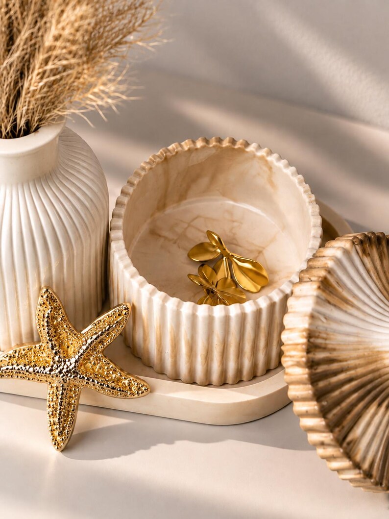 May include: A round, ribbed, cream-colored container with a marble-like finish, containing two gold-toned leaf-shaped ornaments. Beside it sits a gold-toned starfish, a matching ribbed lid, and a tall, ribbed, cream-colored vase with dried grass.