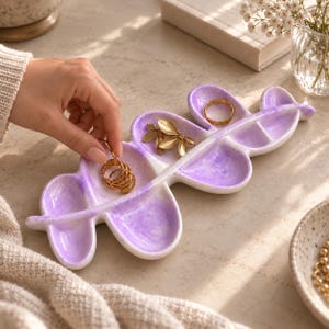 May include: A leaf-shaped ceramic trinket tray featuring a marbled purple and white finish with multiple rounded compartments. The tray holds a gold-toned ring and a gold-toned leaf-shaped accessory, providing a decorative surface for jewelry storage.