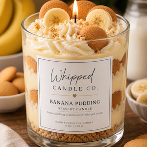 May include: A clear glass jar candle labeled Whipped Candle Co. Banana Pudding Dessert Candle, 9 oz | 255 g. The candle features layers of yellow pudding, round vanilla wafers, and white whipped cream topping with a lit wick in the center.