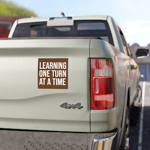 Op de afbeelding: Een vierkante bruine bumpersticker op de achterklep van een lichtgekleurde pick-up truck. De sticker bevat witte schreefloze tekst met de tekst "LEARNING ONE TURN AT A TIME", gerangschikt in vier regels tegen de effen bruine achtergrond.