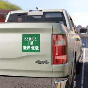 Peut inclure: Un autocollant carré vert avec le texte blanc BE NICE, I'M NEW HERE, apposé sur le hayon d'un pick-up de couleur claire. L'autocollant est placé sur le côté gauche de la benne du camion, près du feu arrière vertical de couleur rouge.