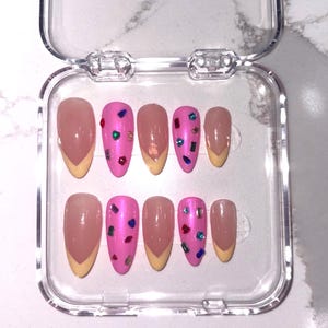 Peut inclure: Un ensemble de dix faux ongles dans un boîtier de rangement en plastique transparent. Les ongles présentent un mélange de rose uni avec des ornements en pierres précieuses colorées et des pointes françaises nude à bords jaune clair.