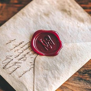 May include: A vintage-style cream-colored paper envelope featuring handwritten cursive script in black ink. The envelope is sealed with a circular, deep red wax seal that displays an embossed letter design in the center of the wax surface.