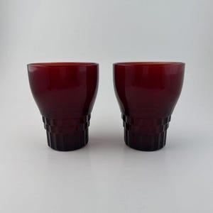 May include: Two matching deep red glass tumblers featuring a smooth, rounded upper rim that transitions into a faceted, geometric base with vertical ridges. The glassware has a translucent finish and a wide, open mouth design.