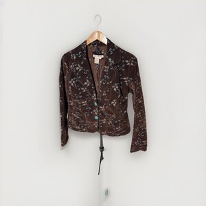 May include: A dark brown velvet blazer featuring a delicate embroidered pattern of light blue and white floral vines. The jacket has a notched collar, long sleeves, and two turquoise-colored buttons down the front. A label reads P.A.M.E.R.
