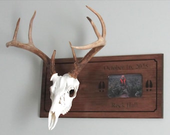 May include: A white deer skull with brown antlers mounted on a rectangular dark wood plaque. The plaque features the engraved text October 16, 2025 and Rock Hall, two deer hoof prints, and a rectangular photo of a person in orange gear.
