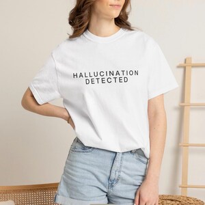 May include: A white short-sleeved crew neck t-shirt featuring the black text HALLUCINATION DETECTED printed in a clean sans-serif font across the chest area. The garment is paired with light blue denim shorts with a cuffed hemline.