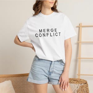 May include: A white short-sleeve t-shirt featuring the black text MERGE CONFLICT printed in a sans-serif font across the chest, paired with light blue denim shorts that have a folded hemline at the bottom of each leg.