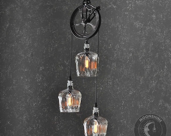 Op de afbeelding: Een zwart metalen katrolsysteem met drie glazen fles hanglampen. De flessen zijn helder en hebben een getextureerd oppervlak. De lampen hangen aan het plafond en zijn verlicht. De tekst "Moonshine Lamp Co." is zichtbaar in de rechterbenedenhoek van de afbeelding.