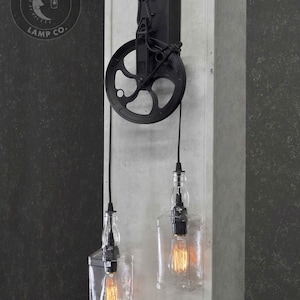 Puede incluir: Aplique de pared de metal negro con polea y dos luces colgantes de botella de vidrio transparente. La aplique tiene una placa de metal negro con un gancho y una polea. Las luces de botella tienen cables negros y bombillas de vidrio transparente.