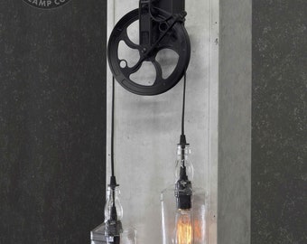 May include: Black metal pulley wall sconce with two clear glass bottle pendant lights. The sconce has a black metal plate with a hook and a pulley wheel. The bottle lights have black cords and clear glass bulbs.