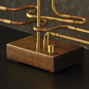 May include: A brass table lamp with a unique design featuring a series of interconnected pipes. The lamp sits on a dark wood base.