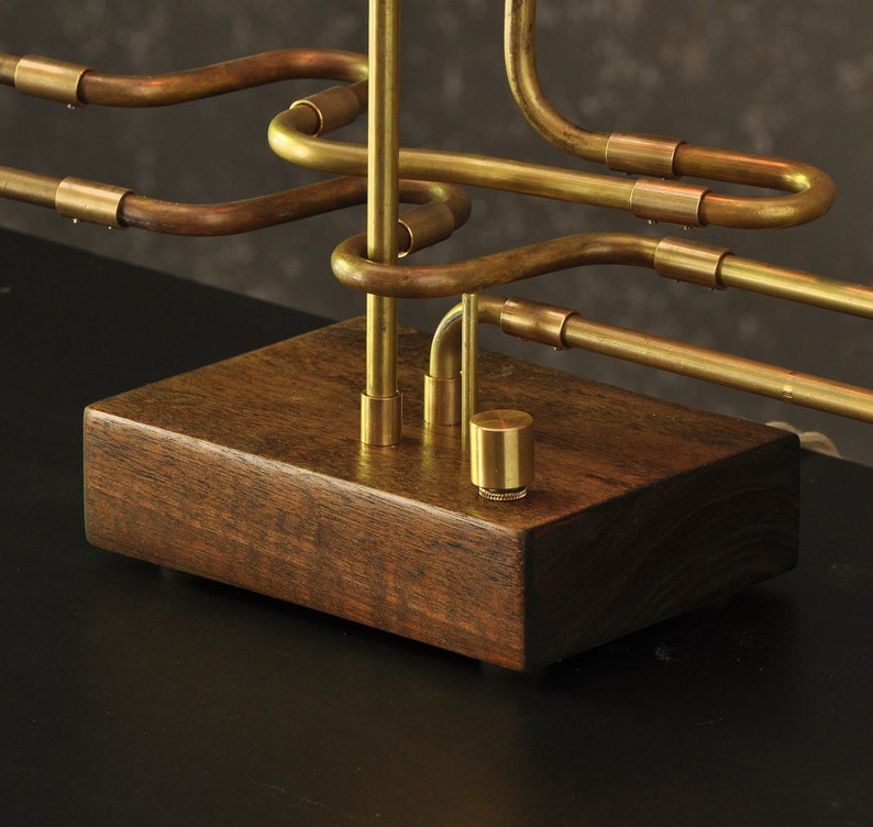 May include: A brass table lamp with a unique design featuring a series of interconnected pipes. The lamp sits on a dark wood base.