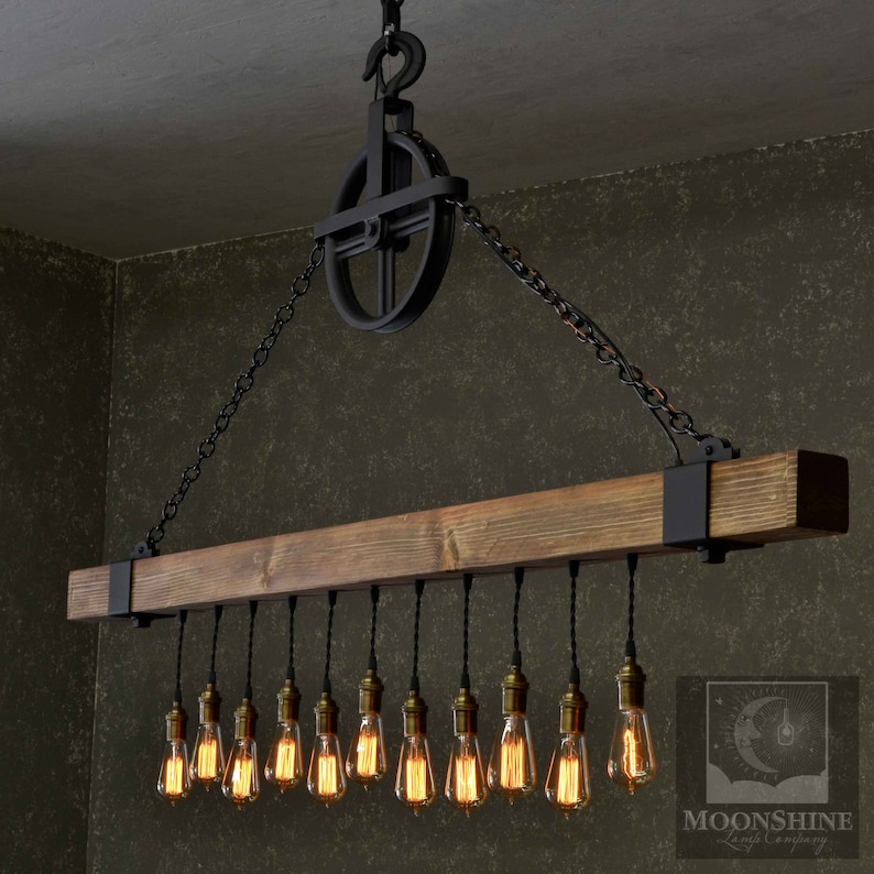 Op de afbeelding: Een rustieke houten balk-kroonluchter met 15 Edison-lampen die aan zwarte snoeren hangen. De kroonluchter hangt aan een zwarte ketting en een katrolsysteem. De balk is donkerbruin gebeitst en heeft aan beide uiteinden zwarte metalen beugels. De lampen zijn allemaal aan en geven een warm licht.