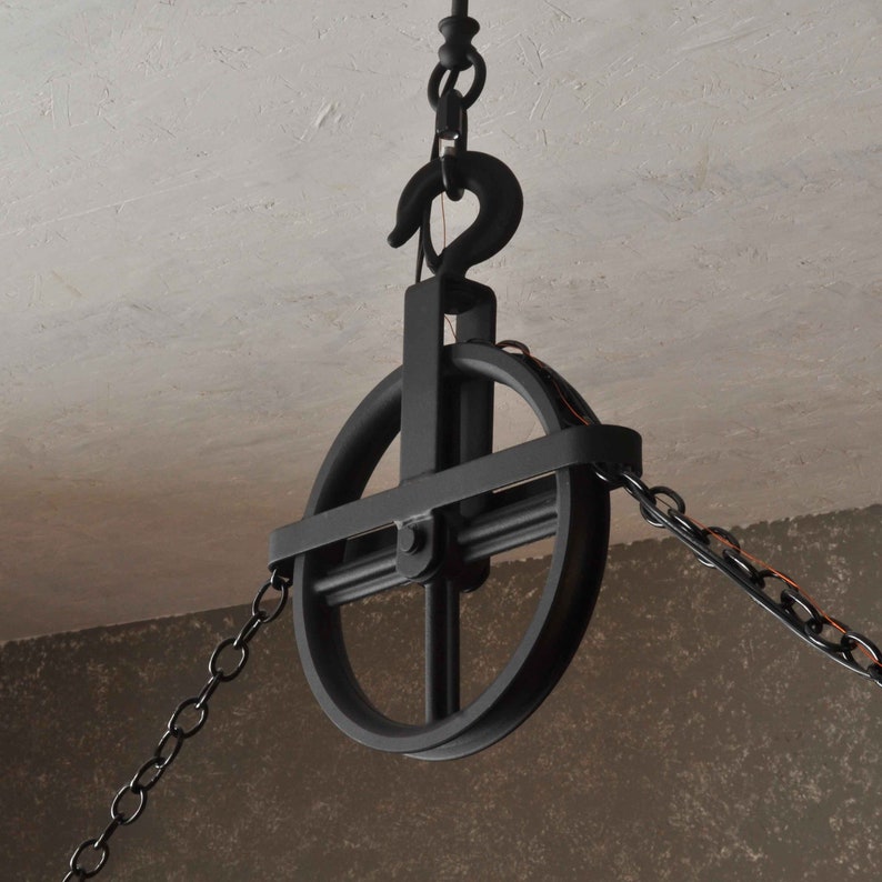 Op de afbeelding: Zwarte metalen katrol met een ketting eraan bevestigd. De katrol heeft een rond wiel met een kruisvormig ontwerp. De ketting is aan de bovenkant van de katrol bevestigd en hangt aan beide kanten naar beneden.