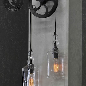 Puede incluir: Dos botellas de vidrio transparente con bombillas colgando de un sistema de poleas negro. Las botellas est&aacute;n suspendidas de cuerdas negras y el sistema de poleas est&aacute; montado en una pared gris. El logotipo "Moonshine Lamp Co." es visible en la esquina superior izquierda de la imagen.