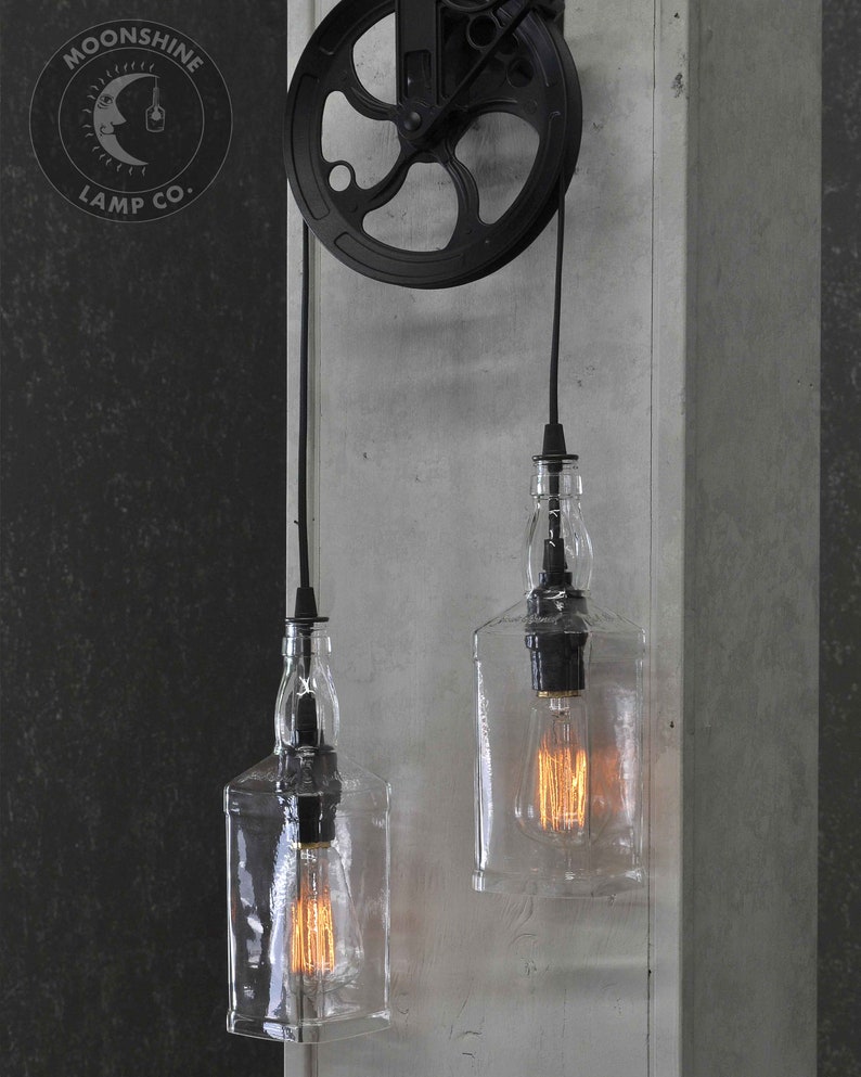 Puede incluir: Dos botellas de vidrio transparente con bombillas colgando de un sistema de poleas negro. Las botellas est&aacute;n suspendidas de cuerdas negras y el sistema de poleas est&aacute; montado en una pared gris. El logotipo "Moonshine Lamp Co." es visible en la esquina superior izquierda de la imagen.