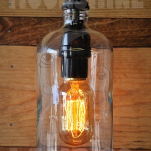 May include: A clear glass bottle pendant light with a black socket and a vintage-style light bulb. The light is hanging from a black cord and is illuminated.