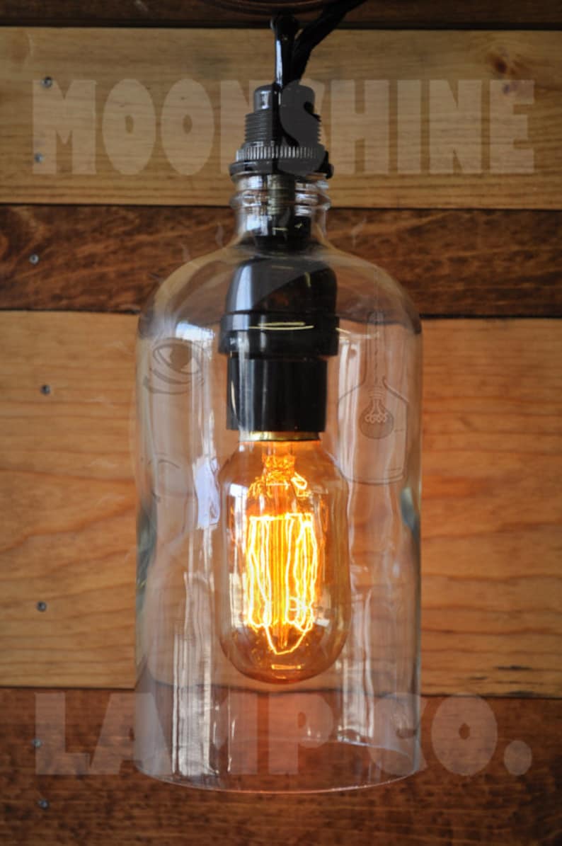 May include: A clear glass bottle pendant light with a black socket and a vintage-style light bulb. The light is hanging from a black cord and is illuminated.