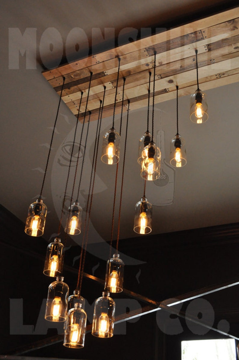 May include: A rustic chandelier made from reclaimed wood with 12 glass bottle light fixtures. The chandelier is hanging from the ceiling and the lights are turned on, casting a warm glow.