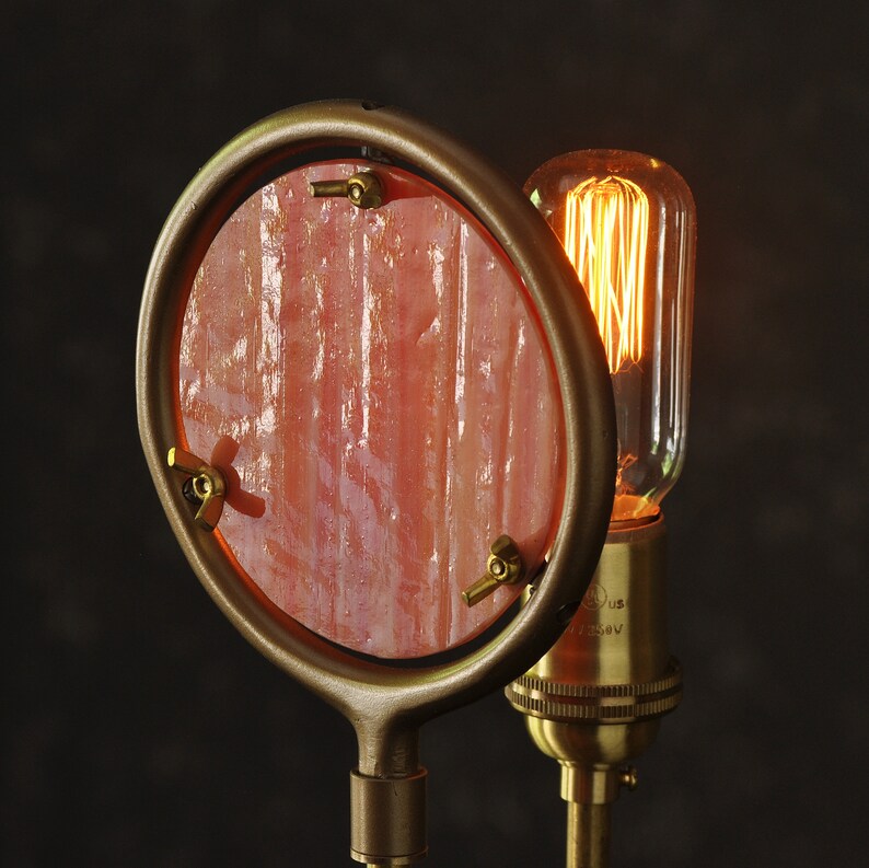 May include: A brass lamp with a pink glass shade and a vintage-style light bulb. The lamp has a round, open-top shade with a decorative design.