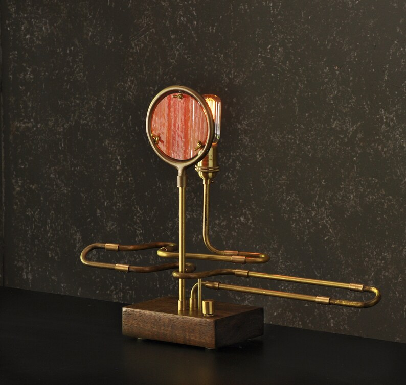 May include: A brass table lamp with a round, red-orange glass shade and a unique, curved design. The lamp has a wooden base and a single light bulb.